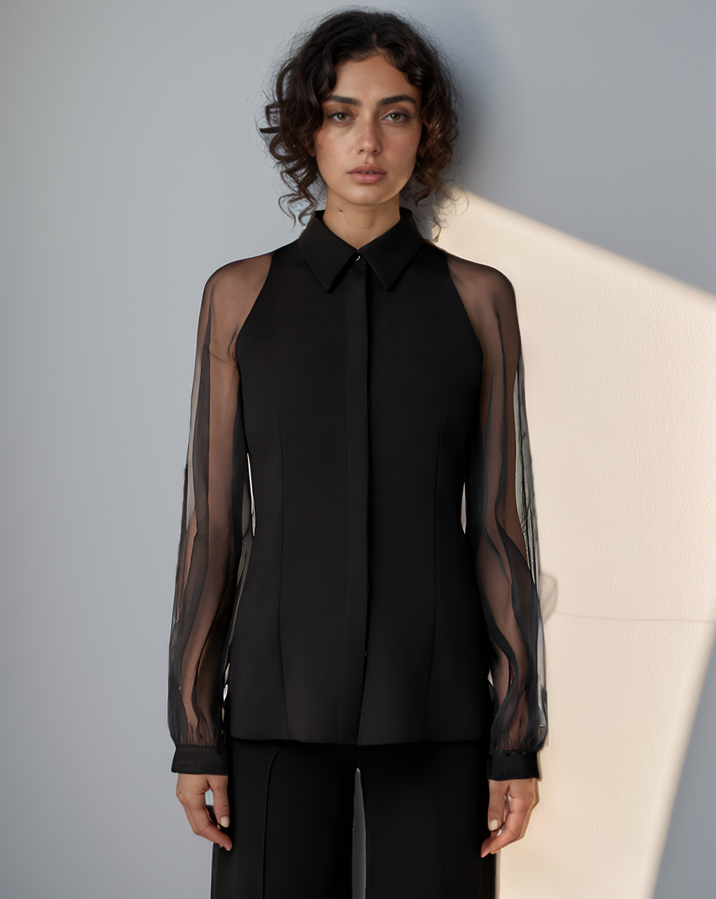 Woman wearing a black sleeveless blouse with sheer sleeves against a neutral background