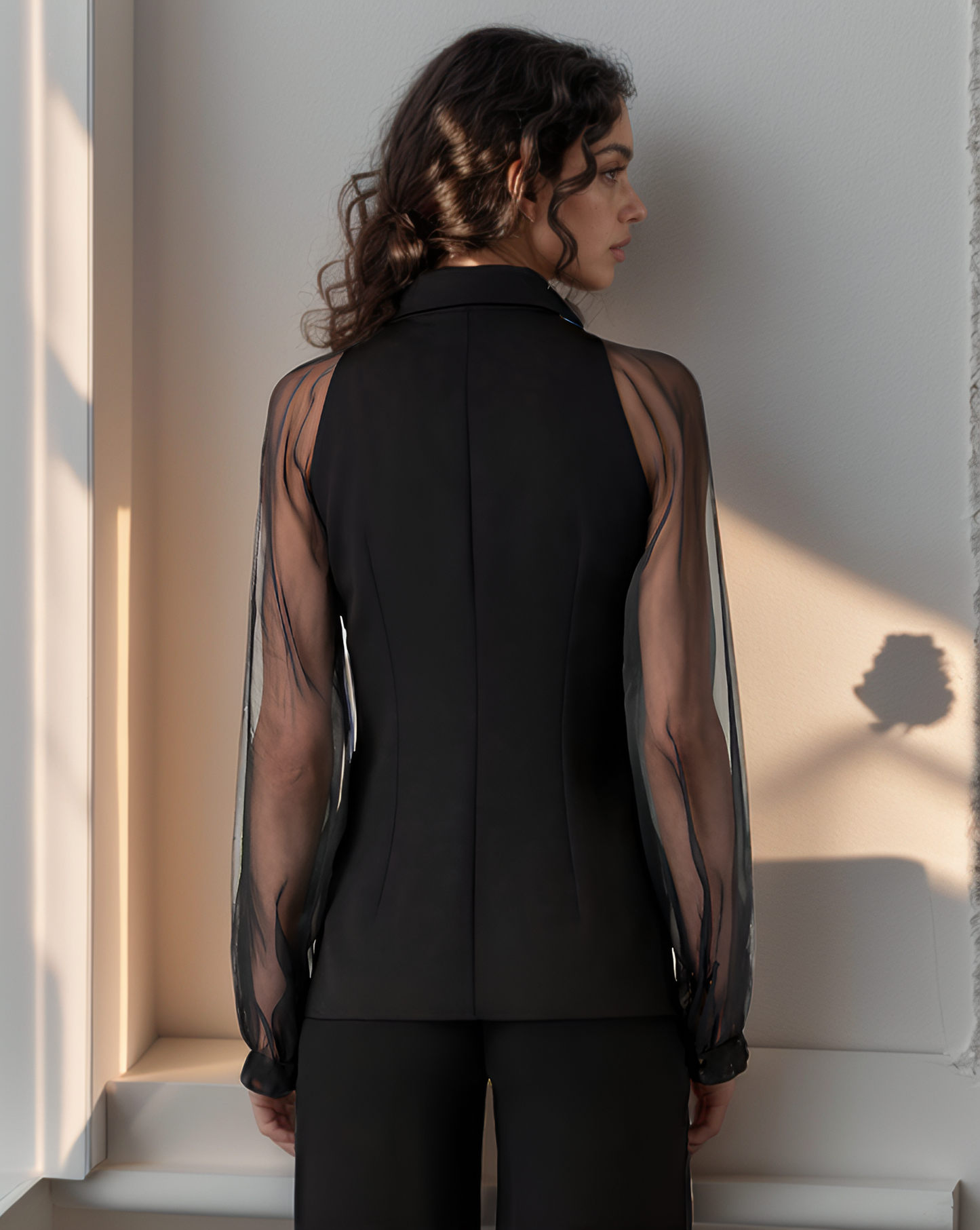 Woman wearing a black outfit with sheer sleeves against a light wall.