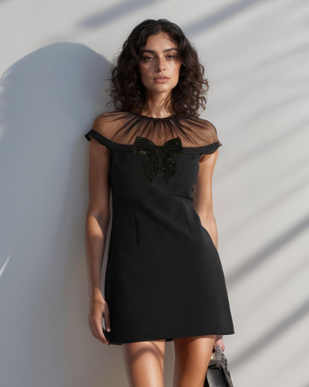 Woman in a black dress standing against a white wall with shadows.
