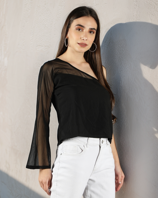 Woman wearing a black top with sheer sleeves and white pants against a plain wall.