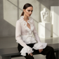 Woman wearing a white blouse with ruffled details sitting on a bench in a modern interior setting.
