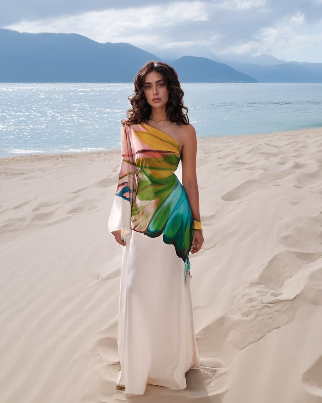 Woman in a colorful dress standing on a sandy beach with mountains and ocean in the background