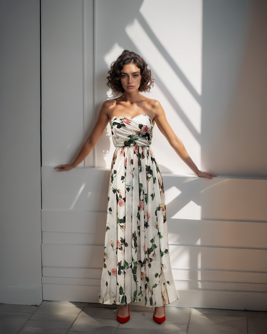 Woman wearing a rose floral strapless dress standing against a white wall with sunlight casting shadows.