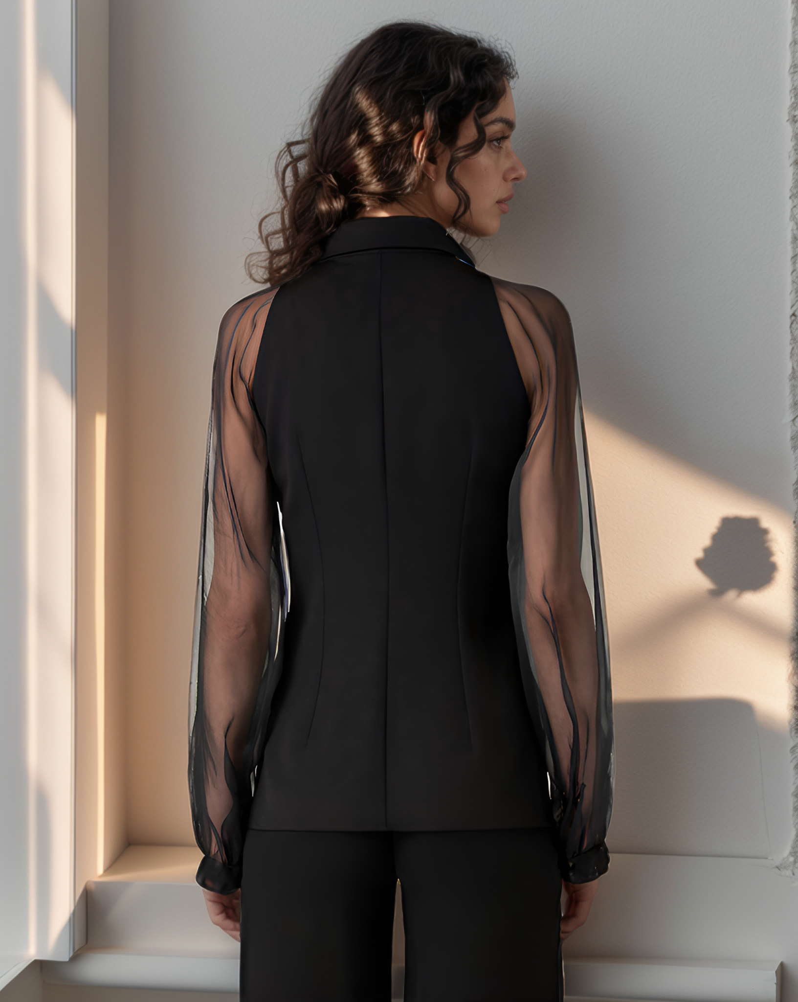 Woman wearing a black outfit with sheer sleeves against a light wall.