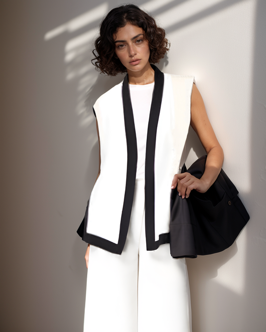 Woman wearing a white vest with black trim and holding a black handbag against a neutral background