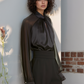 Woman wearing a dark blouse with sheer sleeves against a light wall.