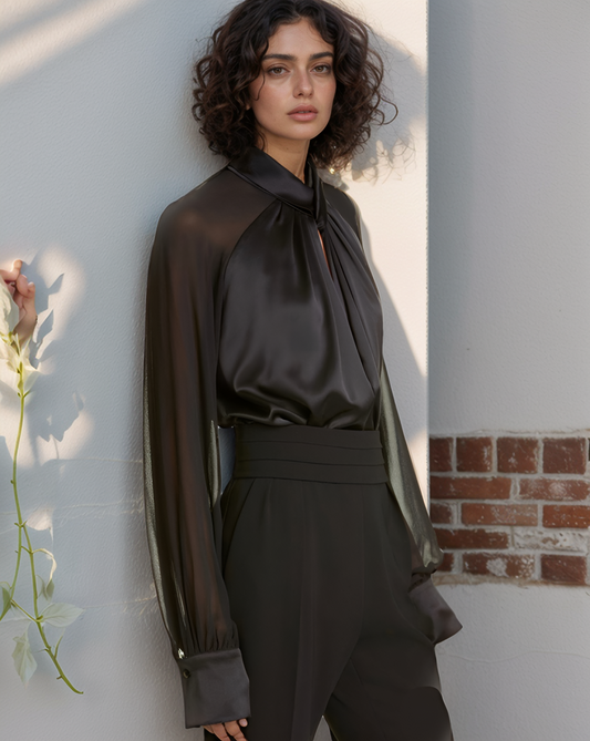 Woman wearing a dark blouse with sheer sleeves against a light wall.