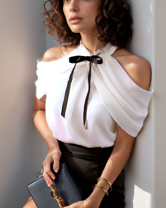 Woman in a white blouse with a black bow tie and black skirt holding a black clutch against a light gray wall.