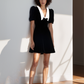 Woman wearing a black dress with a white collar against a white wall.