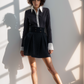 Woman in a black outfit with a white shirt, standing against a light wall.