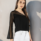 Woman wearing a black top with sheer sleeves and white pants against a plain wall.