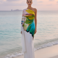 Woman in a colorful one-shoulder dress standing on a beach with ocean and sky in the background