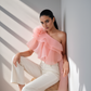 Woman wearing a pink one-shoulder ruffled top and white pants, sitting against a white wall with sunlight casting shadows.