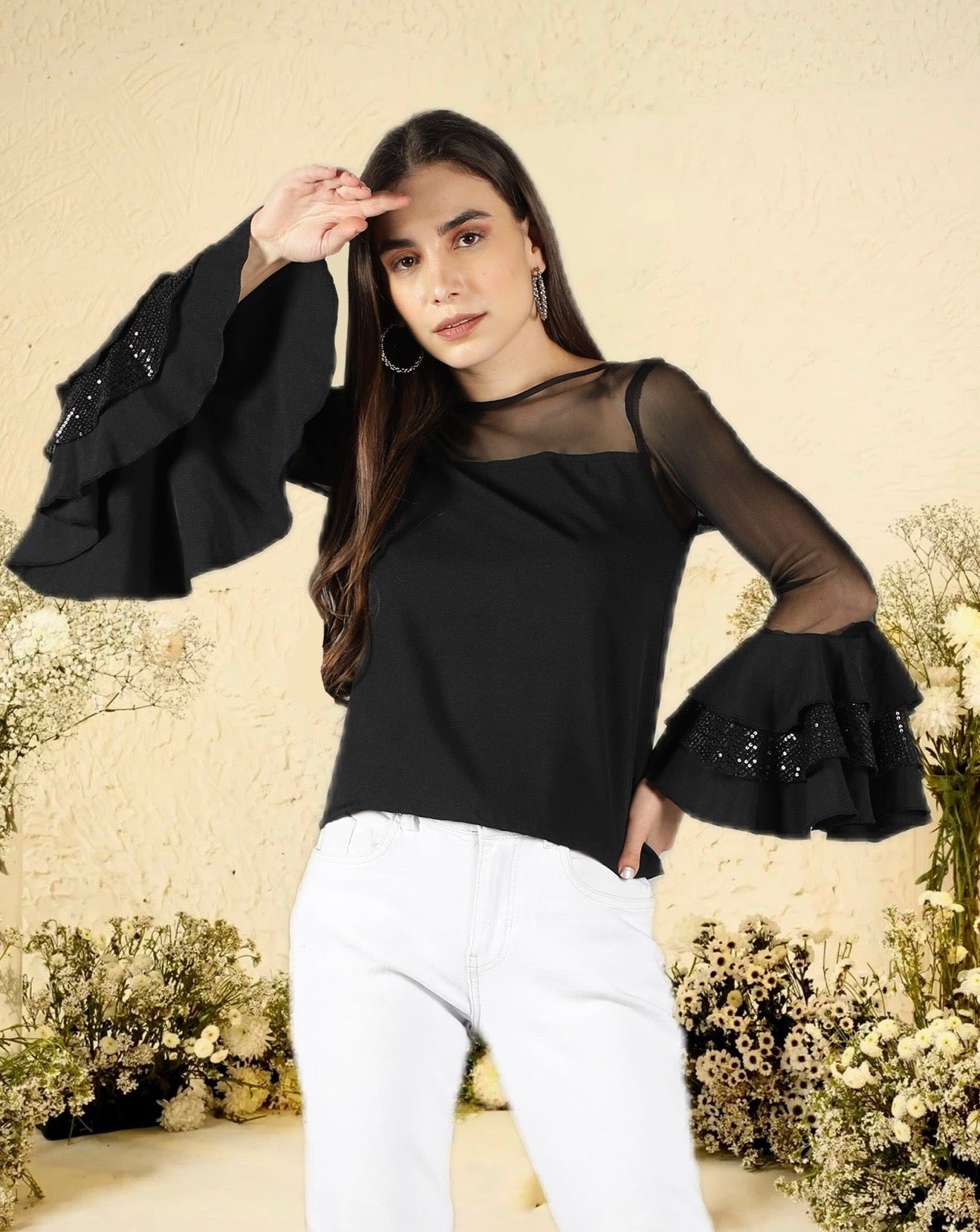 A woman modeling a black top with sequined ruffle sleeves and a round neckline, paired with white pants.